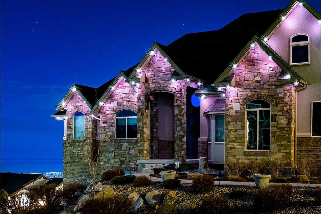 Stone home at dusk with pink and white downlights along the roofline